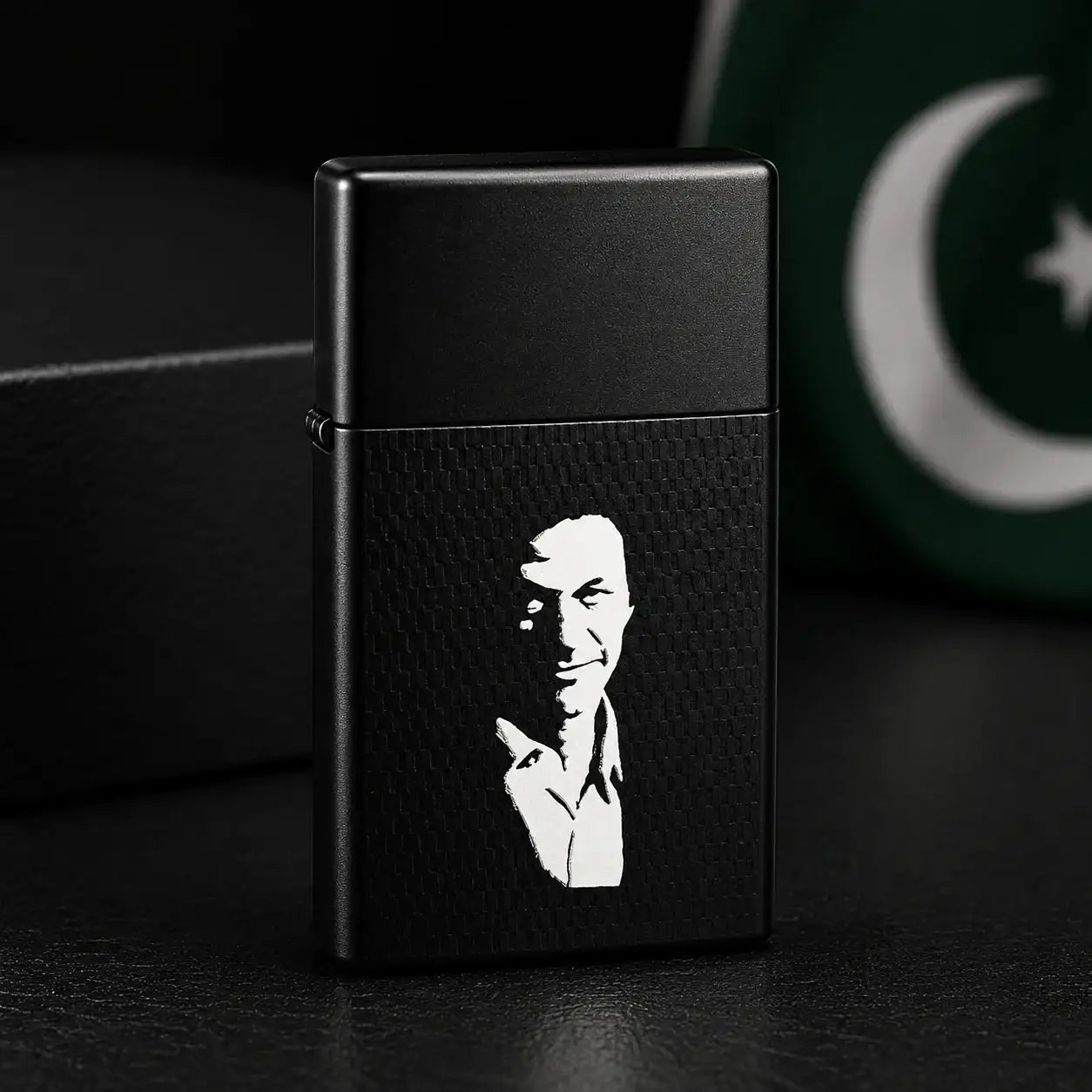 A matte black lighter with a carbon-fiber-textured front panel featuring a white graphic portrait, standing on a dark surface with a blurred green and white flag in the background.