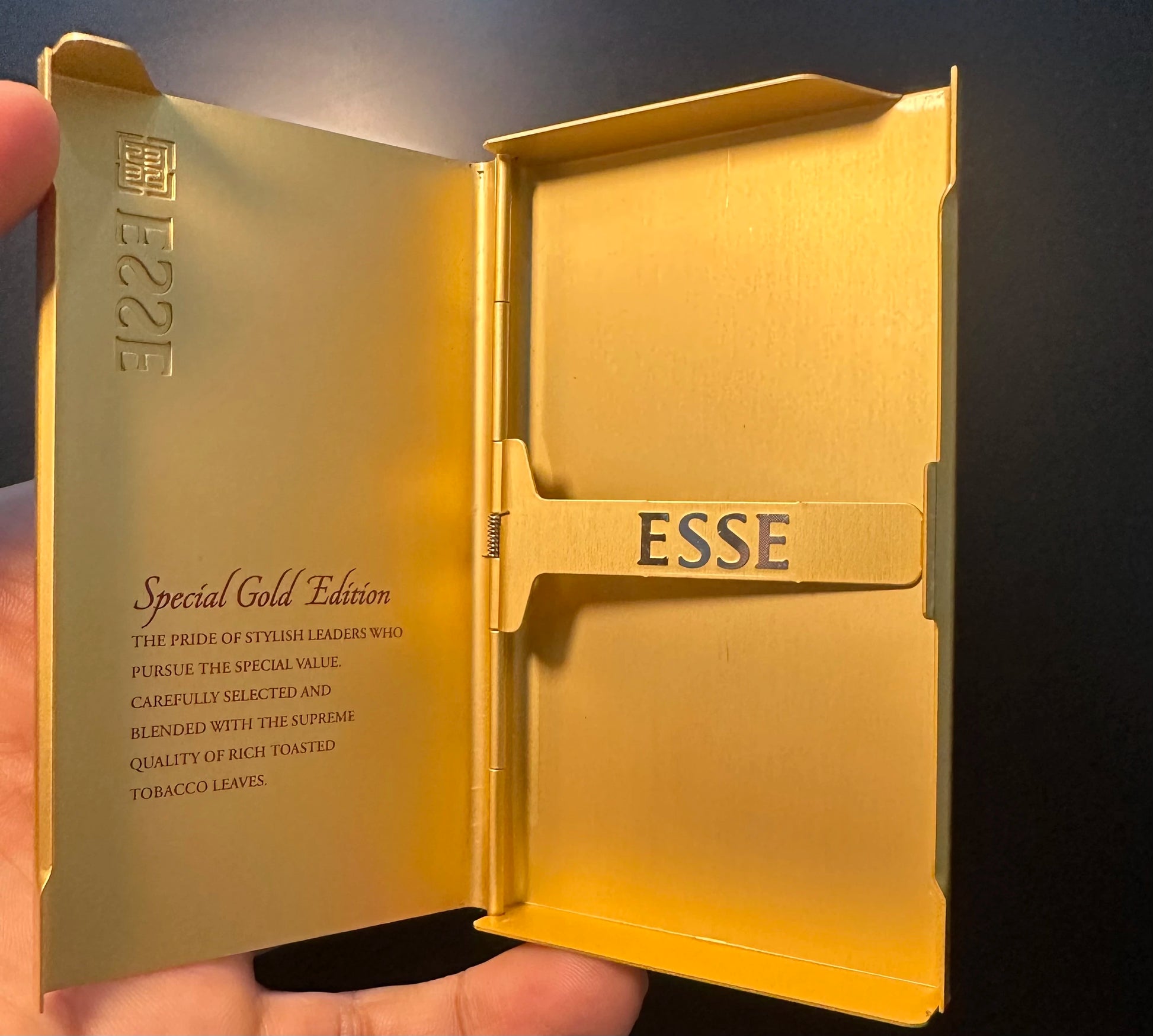 Gold ESSE cigarette case held open by a hand against a dark background