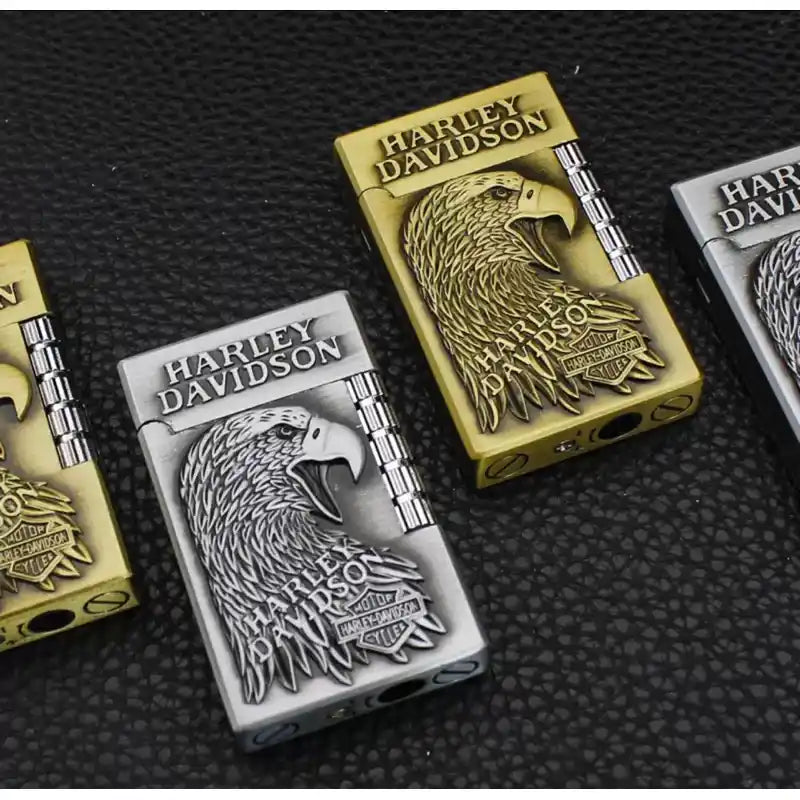 Two Harley-Davidson lighter-style cases shown on a dark textured surface, one in gold-toned metal and one in silver-toned metal, both featuring an embossed eagle head and Harley-Davidson branding.