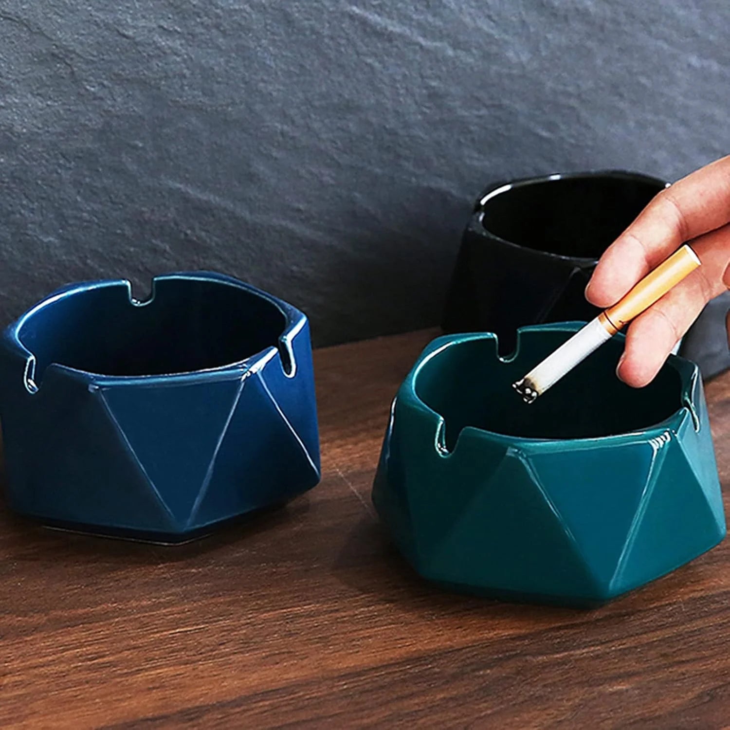 Three geometric ashtrays on a wooden surface with a hand holding a cigarette.