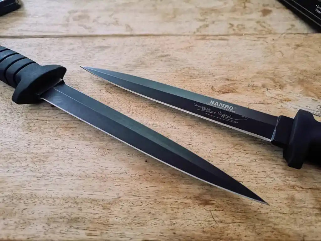 Two knives with black handles on a wooden surface, one branded 'Rambo'.