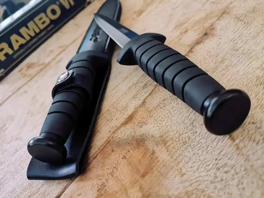 Black knife with textured handle on a wooden surface, with 'Rambo' book in the background.