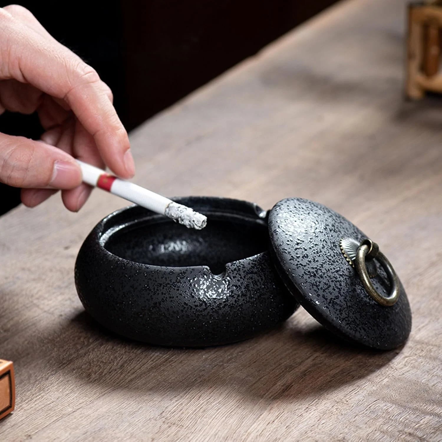 Person placing a cigarette into a black ceramic ashtray on a wooden surface